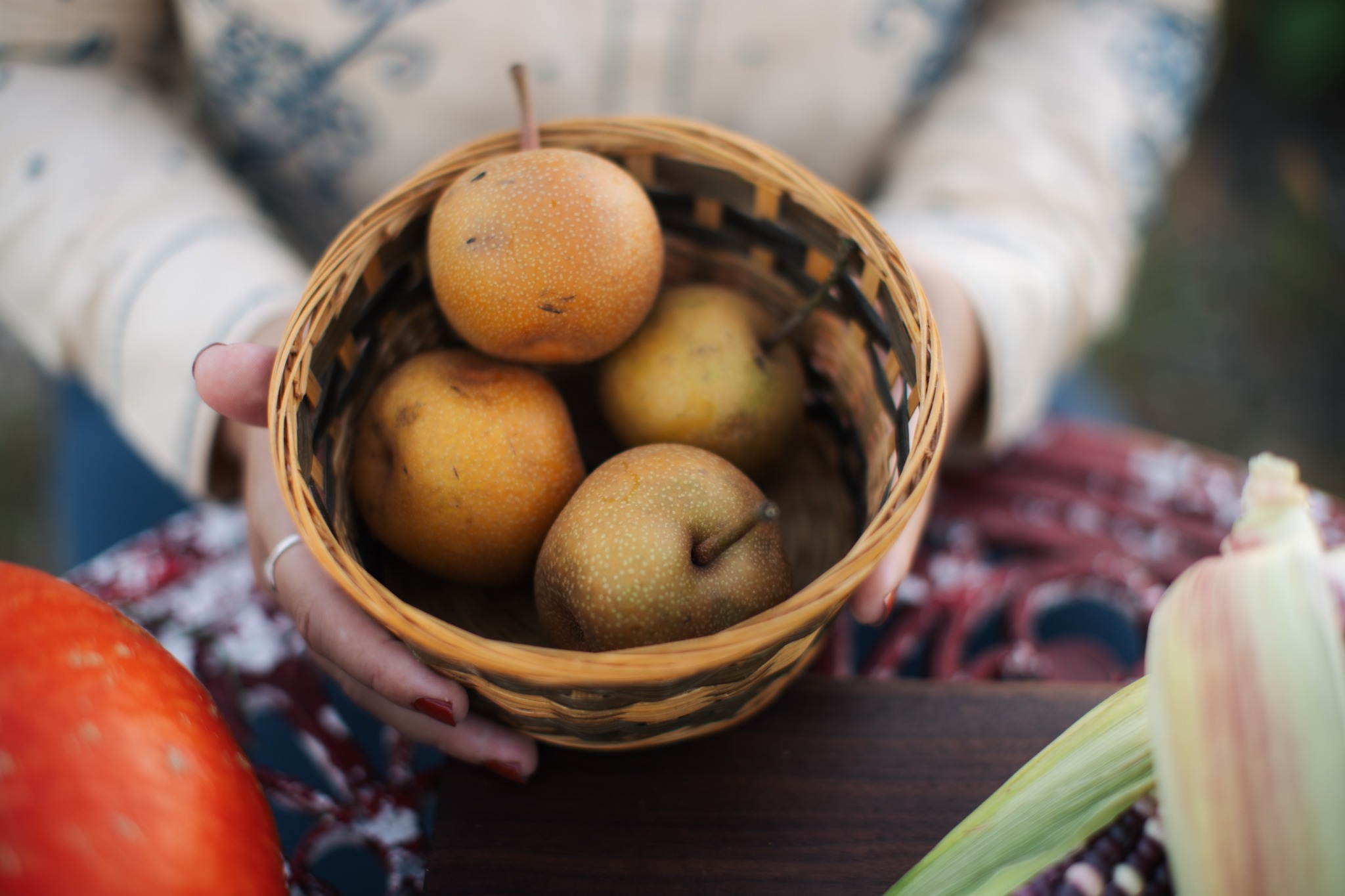 Emma holding a woven basket of pears, with squash and corn on the table.
