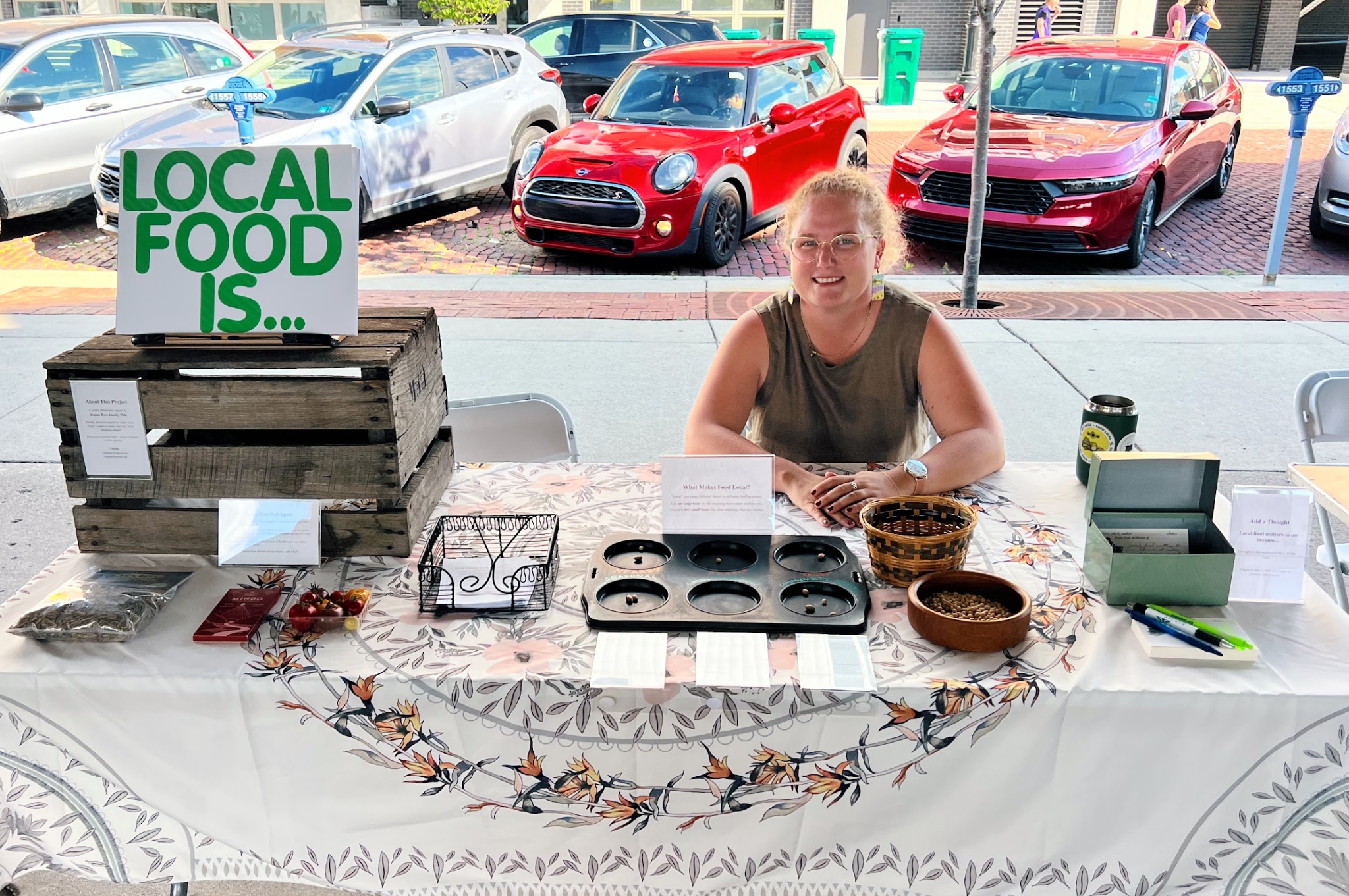 Emma at the What Makes Food Local? exhibit, with food examples, a bean-voting tray, and response cards on the table.